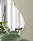 Modern interior with a soft green wallpaper featuring a delicate botanical leaf pattern, partially reflected in a curved mirror, surrounded by lush greenery.