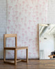 Feminine wallpaper with delicate pink hand-drawn bows on a white background, styled with a natural wood chair and a large mirror reflecting an industrial-style interior.
