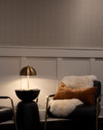Cozy seating area with taupe-toned checkered wallpaper, soft lighting from a brass lamp, and a black leather chair adorned with a shearling throw and rust-colored pillow.