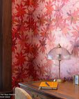 Vibrant red floral wallpaper with large overlapping leaves, creating a warm, textured effect; a wooden desk with a vintage brass table lamp, yellow notebook, pen, and clock in the foreground.