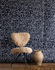 Black wallpaper with intricate white geometric shapes, styled with a beige shearling chair, rustic clay jug, and textured brown rug on wooden floor.