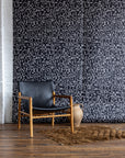 Black wallpaper featuring white abstract geometric patterns, paired with a black leather and wood chair, clay jug, and brown textured rug on wooden floor.