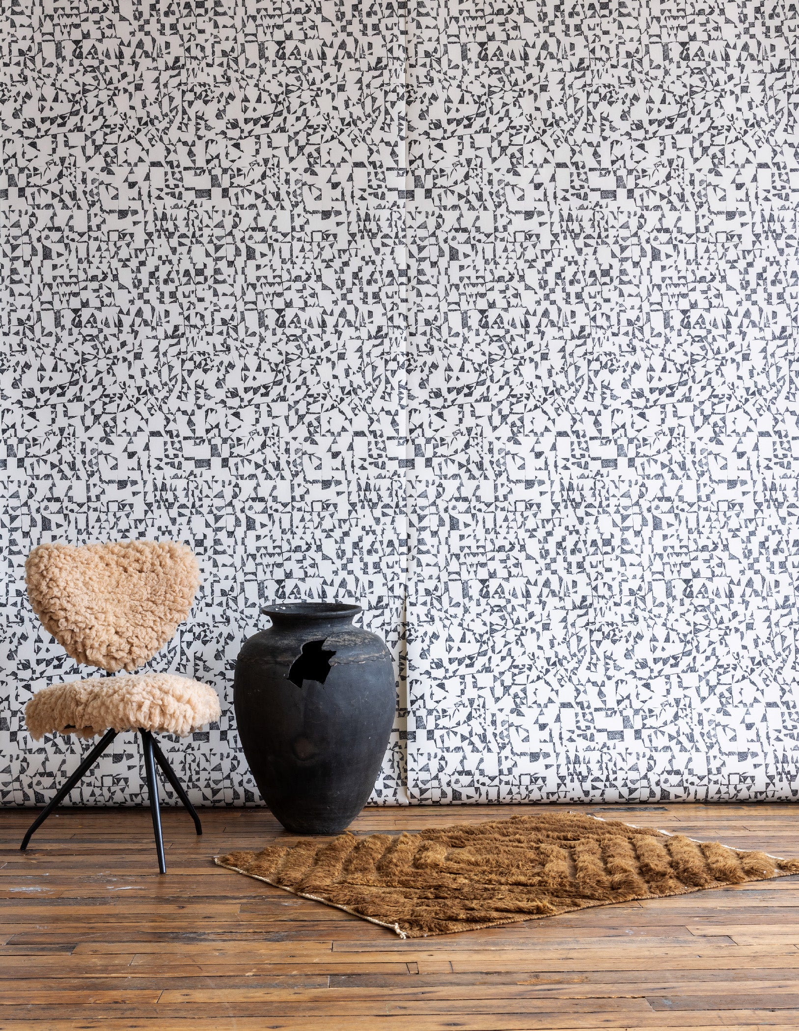 White wallpaper with black abstract geometric pattern, featuring a beige shearling chair, large black clay pot, and brown textured rug on wooden floor.