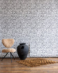 White wallpaper with black abstract geometric pattern, featuring a beige shearling chair, large black clay pot, and brown textured rug on wooden floor.