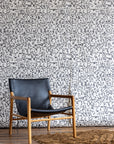 White wallpaper with intricate black geometric shapes, styled with a black leather and wood chair and a brown textured rug on wooden floor.