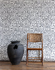 White wallpaper with black geometric pattern, styled with a black clay pot and a woven cane chair on wooden floor.