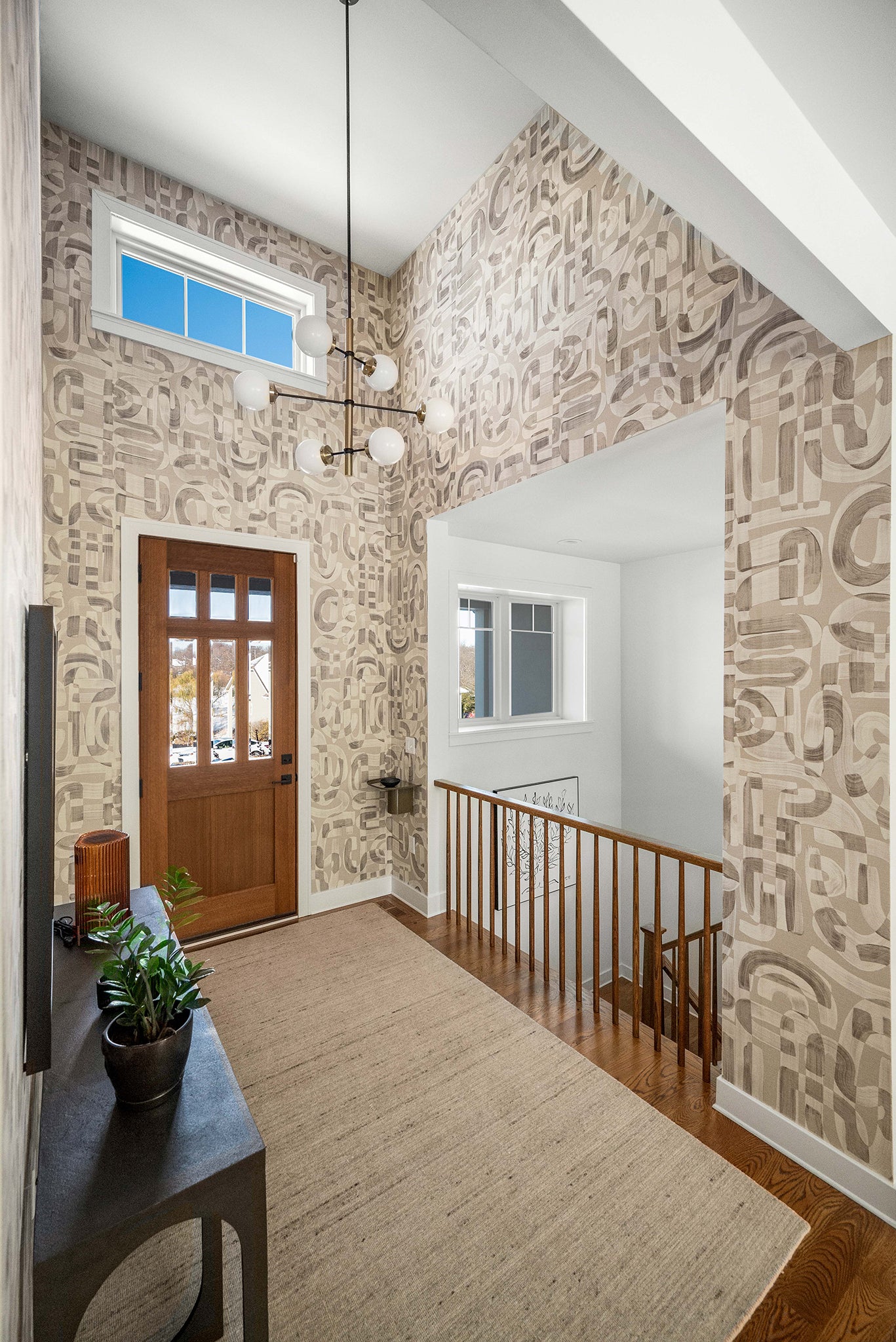 Entryway with neutral-toned grasscloth wallpaper featuring an abstract geometric design, high ceilings, a modern chandelier, and wood accents.