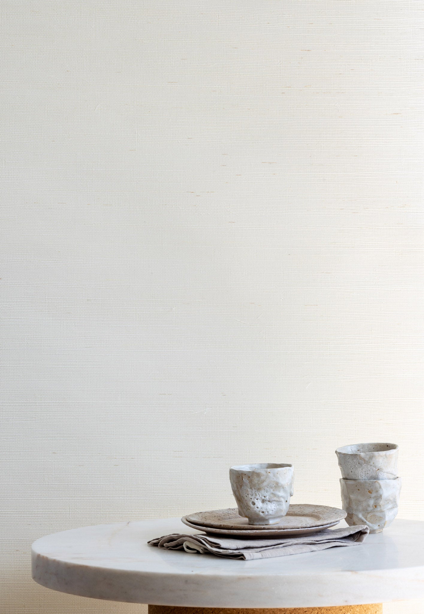 Textured cream grasscloth wallpaper with a natural sisal weave, paired with ceramic tableware on a marble and cork pedestal table.