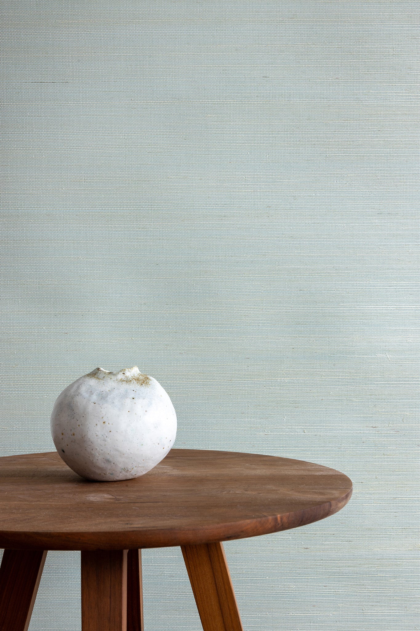 Soft pale blue textured grasscloth wallpaper with a subtle woven finish, styled with a wooden tripod side table and a small white ceramic vase.