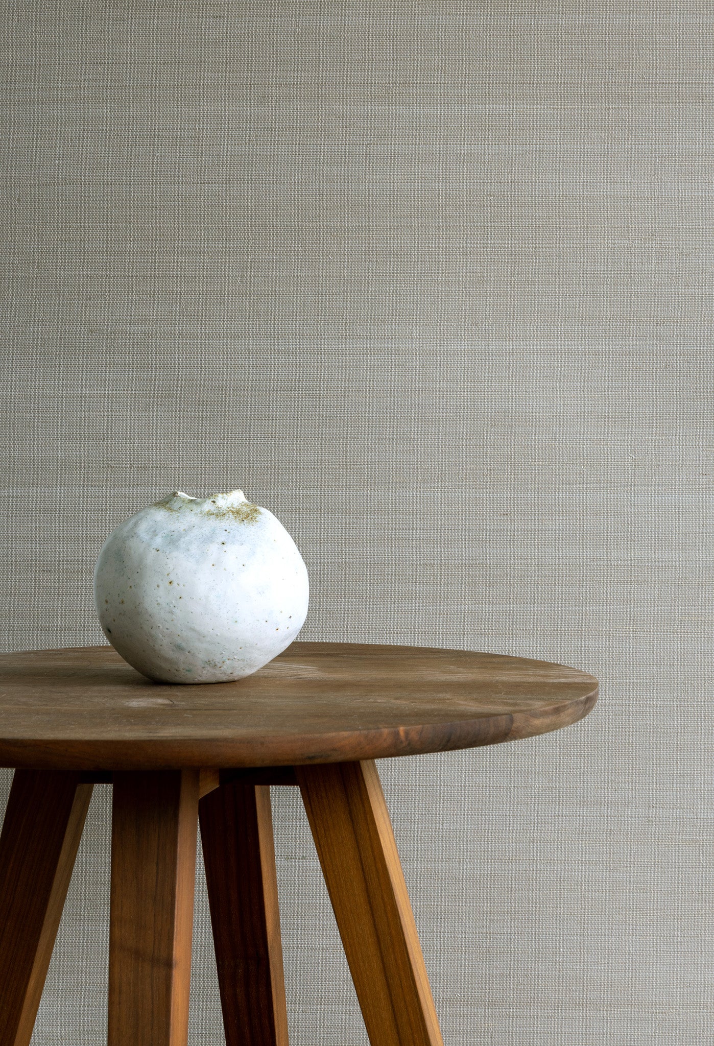 Neutral beige textured grasscloth wallpaper with a subtle woven look, styled with a wooden tripod side table and a small white ceramic vase.