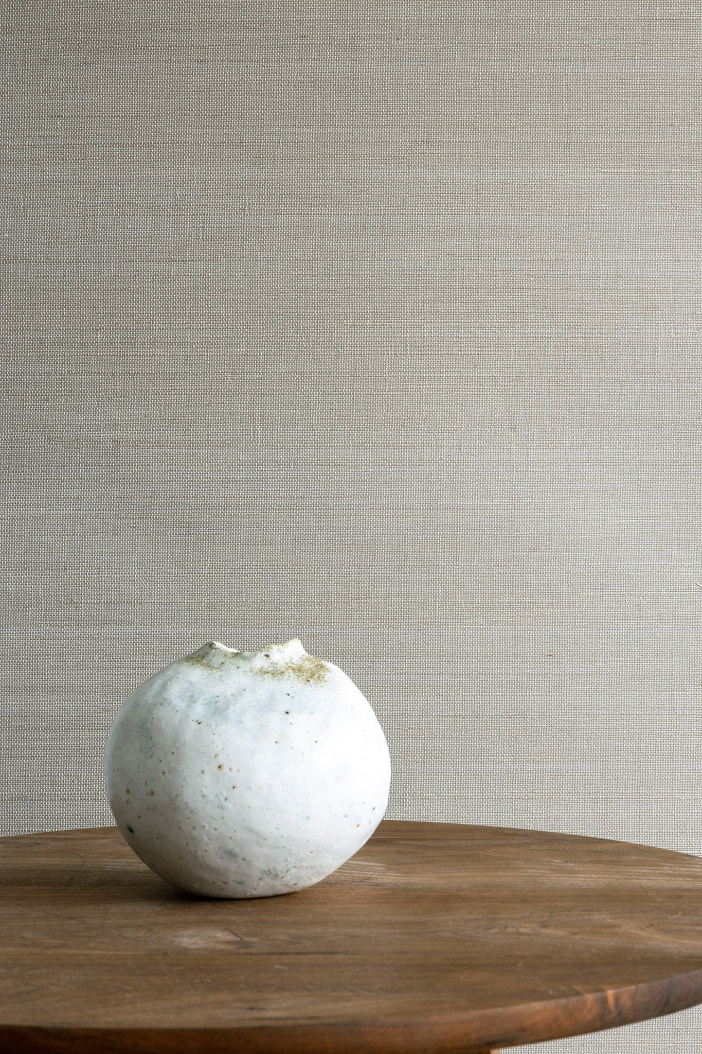 Warm taupe woven-texture grasscloth wallpaper with a minimalist wood side table and a small white ceramic vase in the corner.