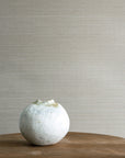 Warm taupe woven-texture grasscloth wallpaper with a minimalist wood side table and a small white ceramic vase in the corner.