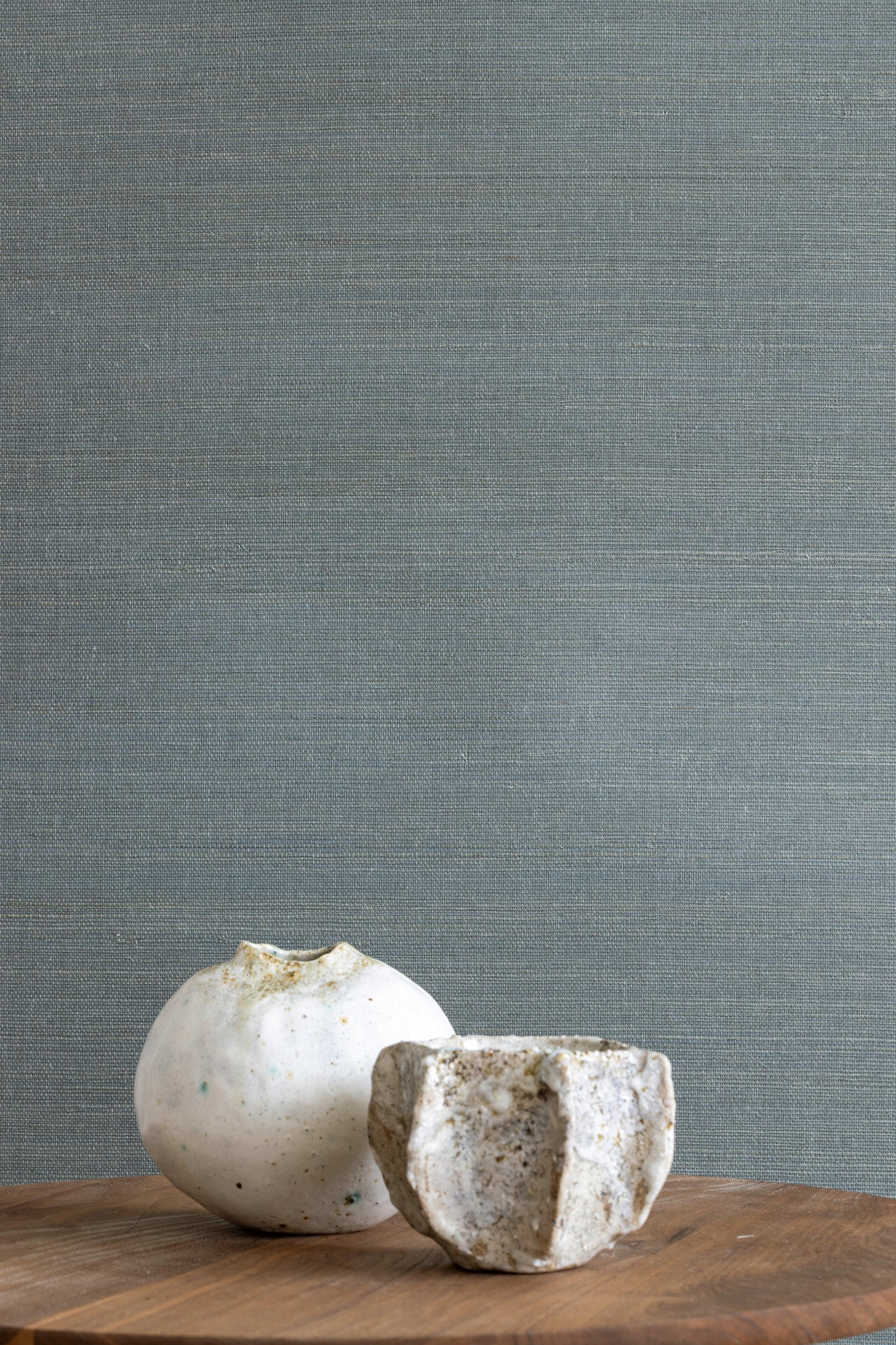 Blue textured grasscloth wallpaper with a subtle woven finish, styled with two white ceramic vases on a wooden surface.