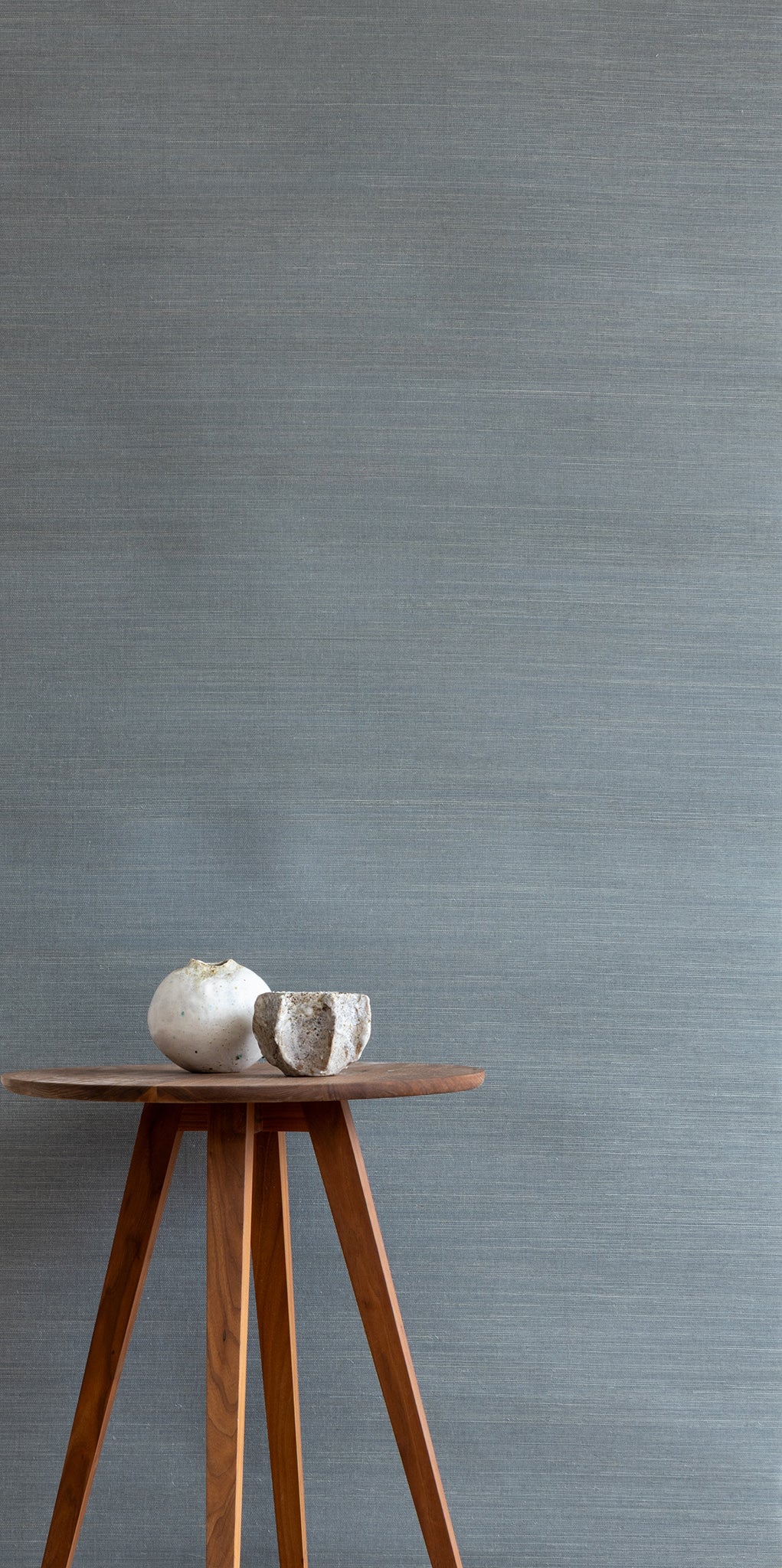 blue textured grasscloth wallpaper with a natural woven look, featuring a wooden tripod side table topped with two white ceramic vases.