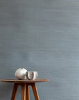 blue textured grasscloth wallpaper with a natural woven look, featuring a wooden tripod side table topped with two white ceramic vases.