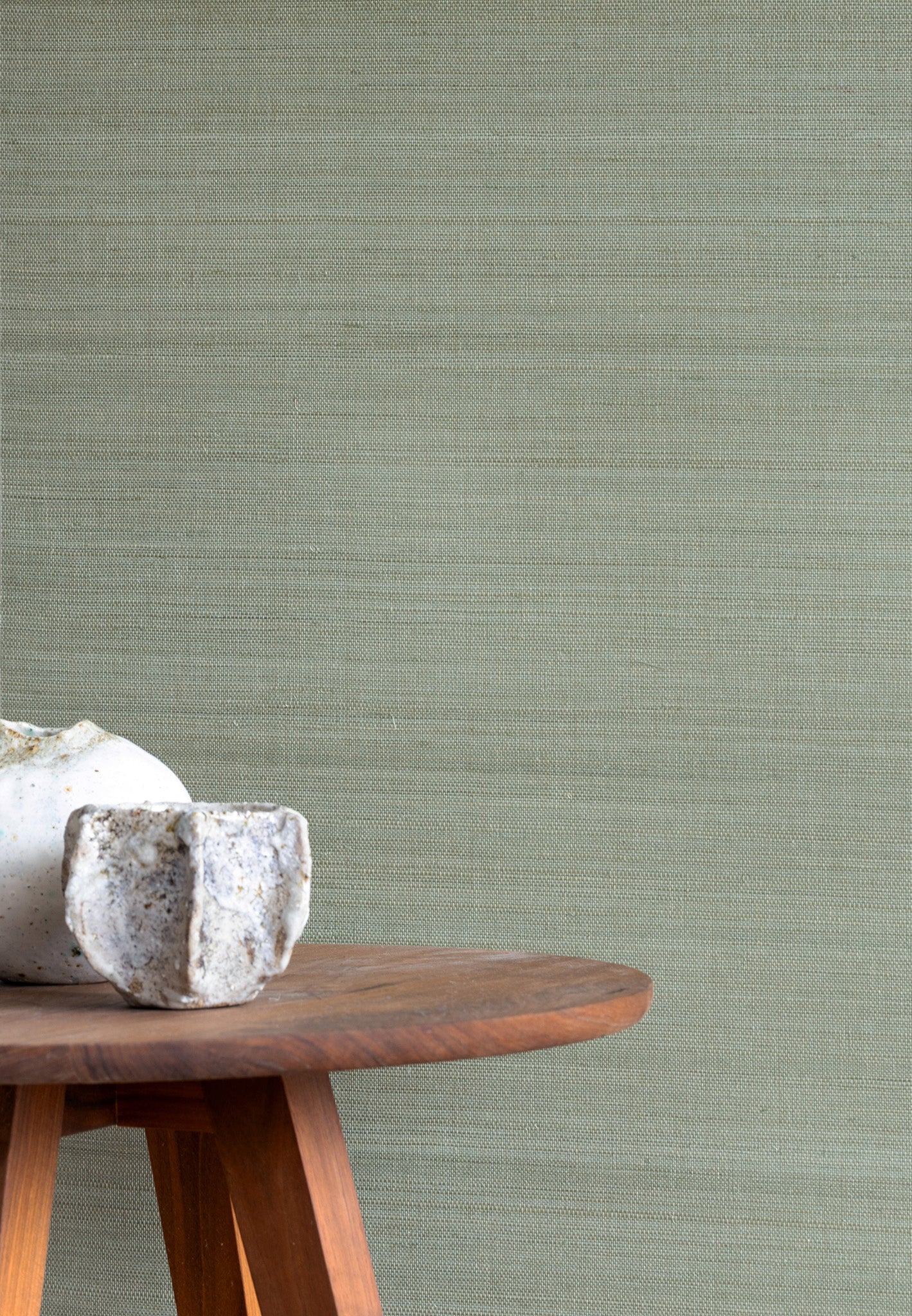 Subtle sage green textured grasscloth wallpaper with a natural woven look, styled with a wooden side table and a small white ceramic vase.