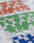 A grasscloth wallpaper sample sheet displaying three color variations of a geometric design—orange, green, and blue—on an off-white background.