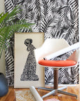 Bold black and white tropical leaf wallpaper with a distorted, ripple-like effect, styled with a modern white chair and vibrant orange cushion.