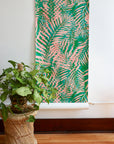 Tropical leaf wallpaper in vivid green and soft coral pink with a ripple effect, partially unrolled against a neutral wall next to a woven plant stand.