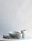Interior setting featuring beige and ivory striped linen wallpaper behind a marble tabletop with ceramic dishware and folded linens.