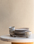 Interior setting with tan and off-white striped linen wallpaper as a backdrop, paired with stacked ceramic dishes and a marble tabletop.