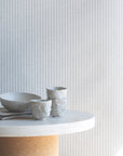 Interior scene with light grey and cream striped wallpaper as a backdrop, paired with a marble table and handmade ceramic cups and bowl.
