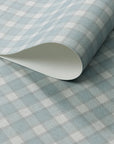 Close-up of a rolled sheet of vintage-style plaid check wallpaper in soft blue and white tones with a linen-textured finish.
