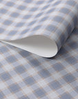 Close-up of vintage plaid check wallpaper in dusty blue and warm beige tones on a soft linen-textured surface, shown with a curled edge to highlight paper quality.