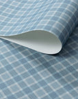 Close-up of vintage plaid check wallpaper in soft blue and ivory tones on a linen-textured surface, with a rolled sheet revealing paper thickness.