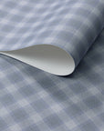Close-up of vintage plaid check wallpaper in cool gray-blue and soft white tones with a linen-textured surface, shown with a rolled edge for detail.