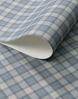 Close-up of vintage plaid check wallpaper in muted denim blue and cream tones with a soft linen texture, shown with a curled edge to display paper thickness.