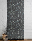 Floral-patterned wallpaper roll against a white brick wall with a vase of branches on a wooden floor.