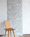 Light botanical wallpaper with a soft blue-gray background and white leafy vine pattern, shown against a white brick wall with a natural wood chair on a neutral woven rug.
