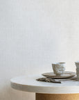 Neutral-toned linen wallpaper with a subtle woven linen texture, styled with ceramic tableware on a marble and cork pedestal table.