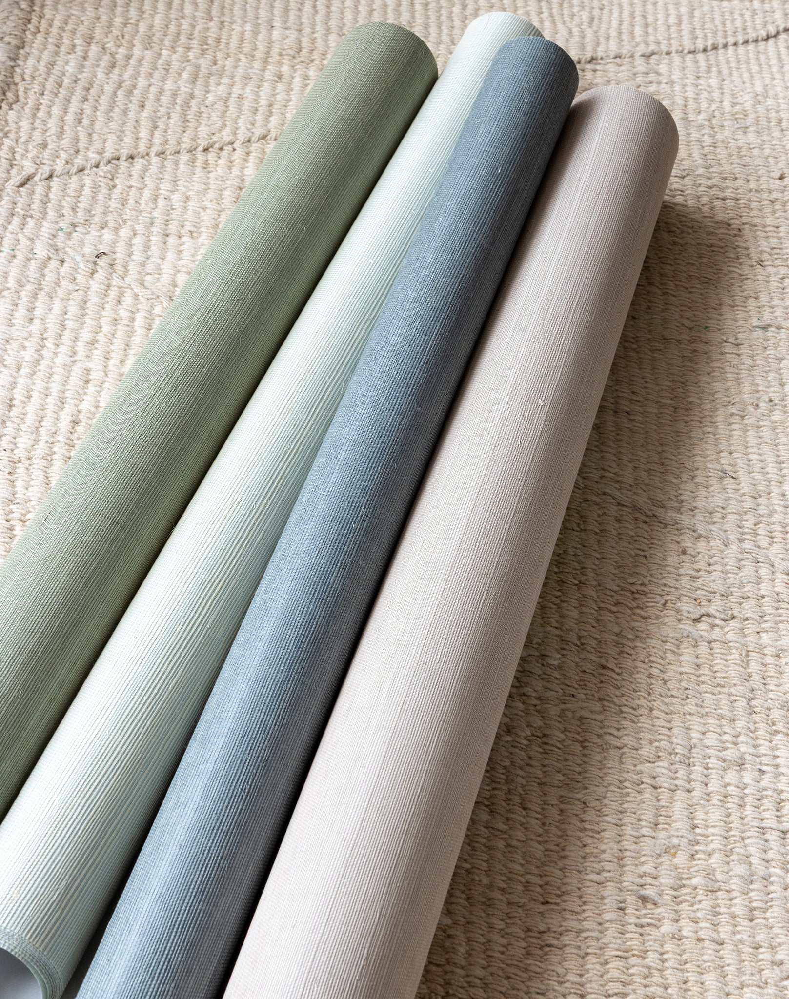 Poppy Neutral-toned grasscloth wallpaper rolls in sage green, light cream, soft blue, and beige, displayed on a woven natural fiber rug.