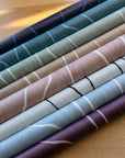 Rolled wallpaper samples in various colors, including navy, teal, green, beige, blush, and plum, all featuring organic vertical line patterns, placed on a light wood surface.