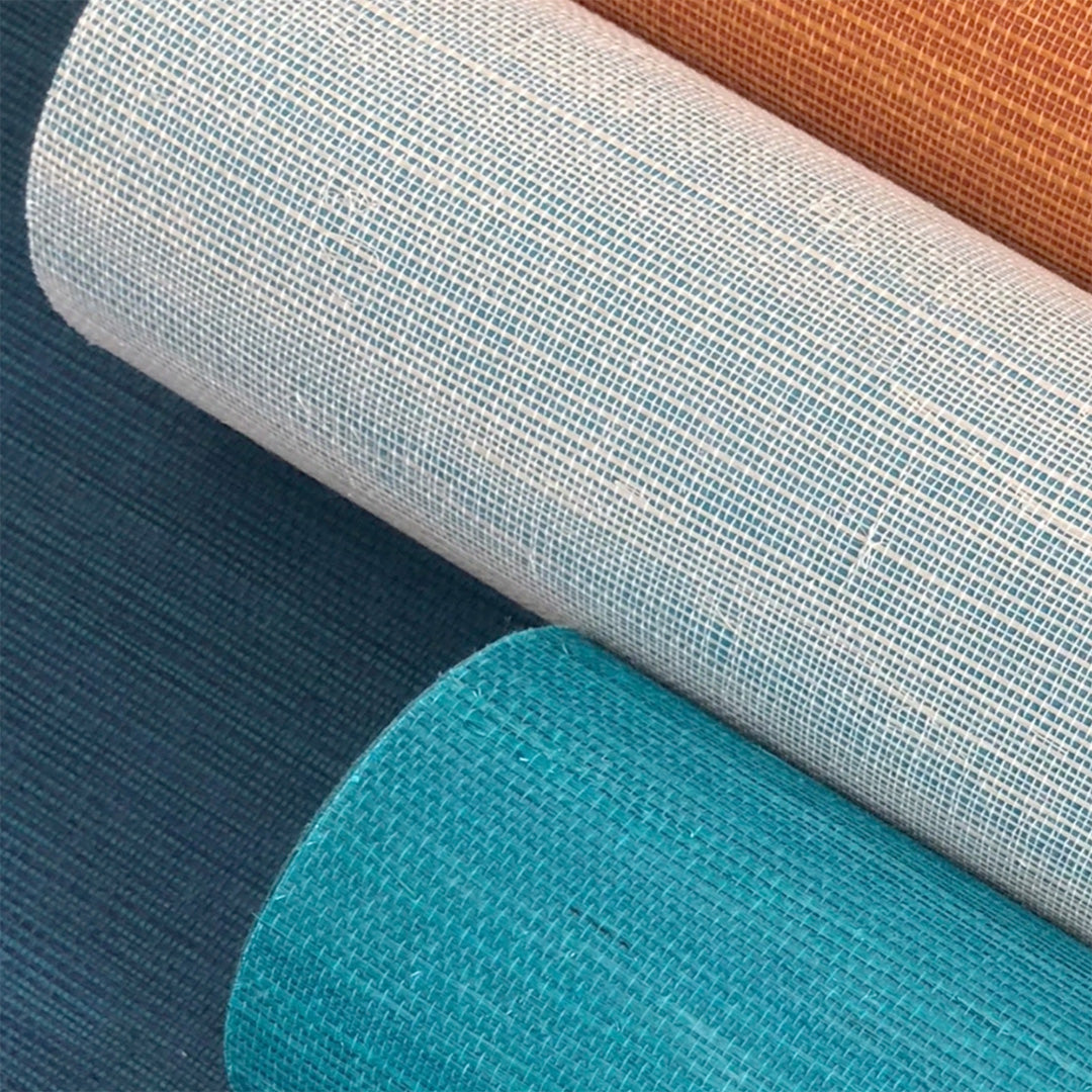 Rolled-up natural fiber grasscloth wallpaper in shades of teal, white, and burnt orange, showcasing a woven grasscloth texture with visible fibers and a grid-like pattern.