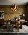 A richly layered dining room showcasing Wave Brass mural wallpaper in deep golden tones, paired with a live-edge wood table, ochre velvet chair, sculptural brass chandelier, dark floral centerpiece, and vintage wood sideboards — Poppy wallpaper