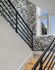 Modern stairwell featuring black-and-white grasscloth wallpaper with an abstract geometric pattern, paired with a black metal railing, light wood steps, and a large window allowing natural light.