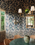 Dining room with blue floral wallpaper covering walls and ceiling, featuring blue leaf patterns; green marble table, wicker chairs, and rustic chandelier with white shades in the foreground.
