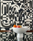 Black and white abstract wallpaper called Down the Rabbit Hole in a dining room setting, with a modern white bowl filled with oranges and an apple placed on a reflective glass table.