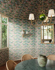 A dining room featuring wallpaper with a lush botanical pattern in green, pink, and blue tones, extending onto the ceiling. The space is decorated with a rope chandelier, wicker chairs, and a green marble table.