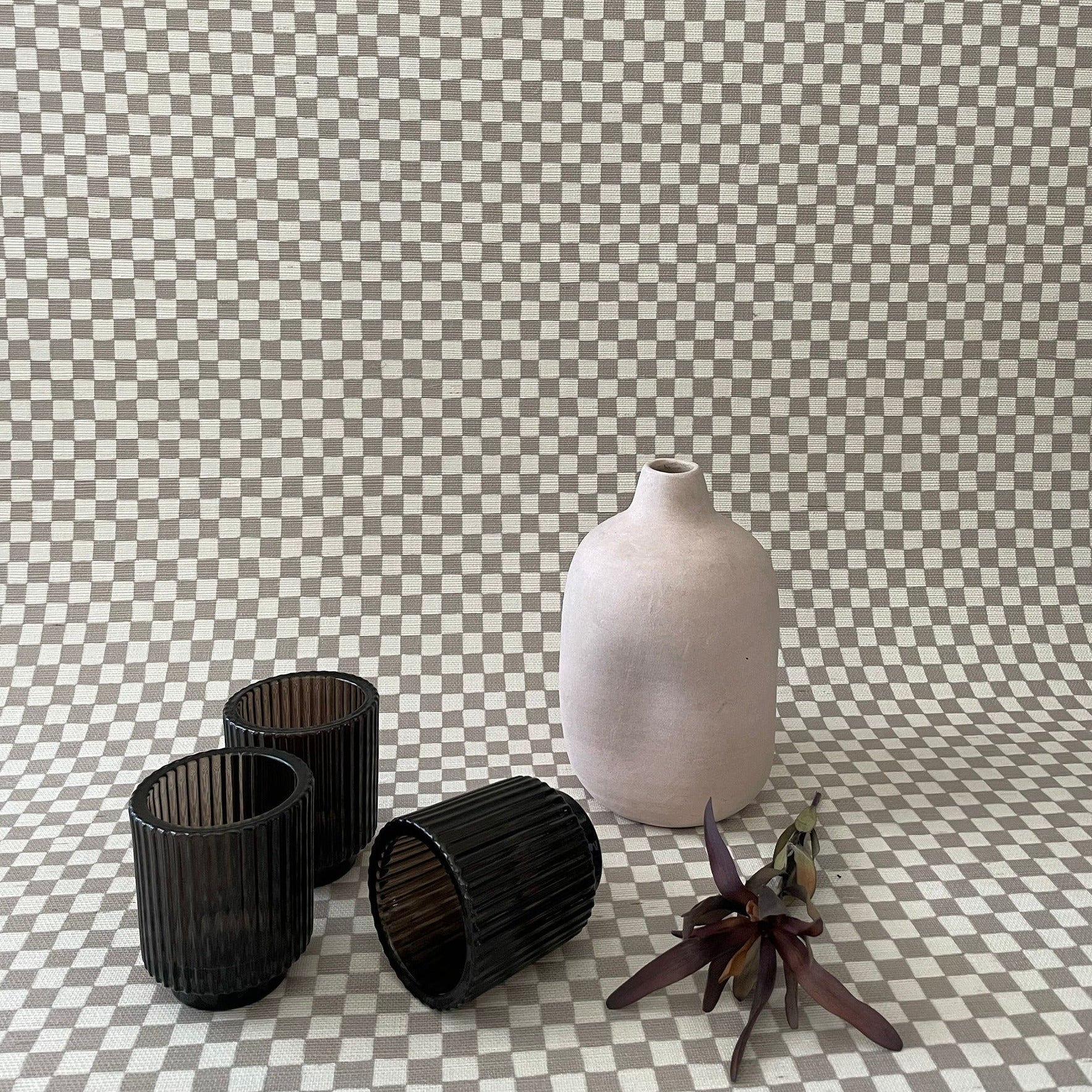 Decorative scene with taupe and white grasscloth checkered wallpaper, featuring black glass candle holders, a matte ceramic vase, and a dried floral arrangement.