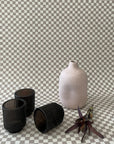Decorative scene with taupe and white grasscloth checkered wallpaper, featuring black glass candle holders, a matte ceramic vase, and a dried floral arrangement.