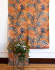 Orange wallpaper with bold floral patterns in muted blue shades, displayed in hanging panels; green leafy plant in a modern metal stand in the foreground.