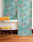 A rolled-out wallpaper featuring a botanical and snake pattern in teal, pink, and gold, displayed against a white wall with a mustard yellow vintage chair.