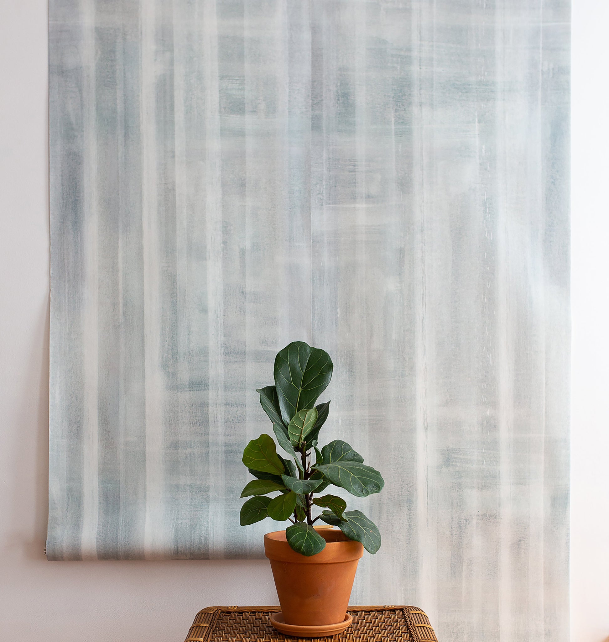 Modern interior with a large hanging panel of gray-toned abstract wallpaper featuring vertical brushstroke stripes, complemented by a potted fiddle-leaf fig on a woven table.