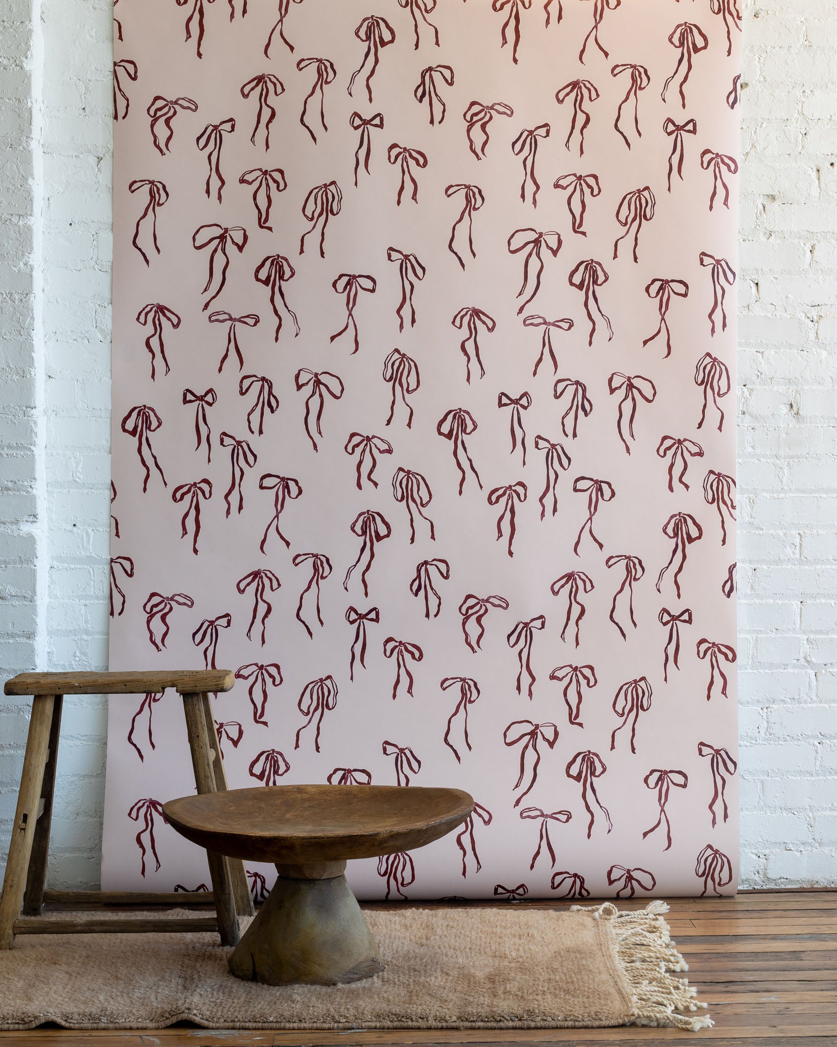 Vintage-inspired wallpaper with hand-drawn red bows on a soft pink background, styled with rustic wooden furniture and a neutral woven rug.