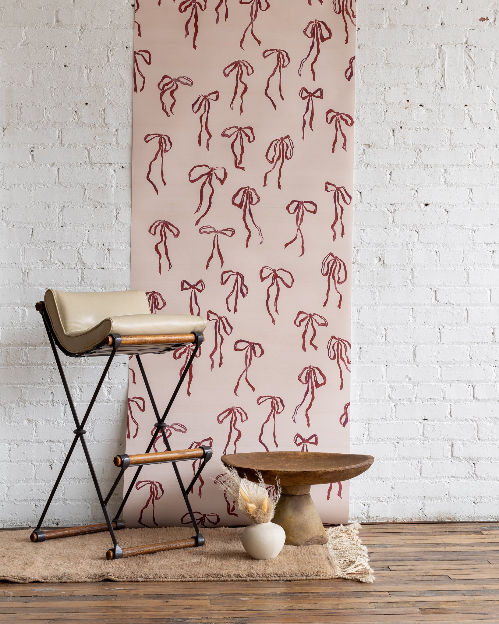 Romantic silk wallpaper with hand-drawn red bows on a soft pink background, styled with a beige barstool, rustic wooden table, and neutral decor.