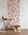 Romantic silk wallpaper with hand-drawn red bows on a soft pink background, styled with a beige barstool, rustic wooden table, and neutral decor.